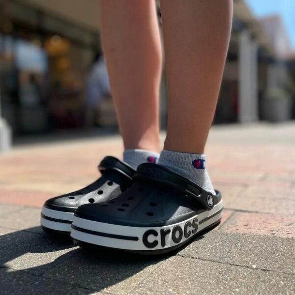 Person wearing black Crocs with a white sole and gray Champion socks standing on a brick-paved sidewalk outdoors.