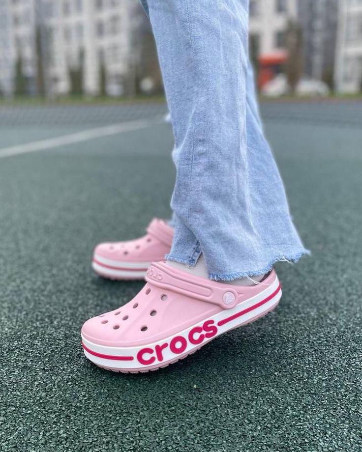 WhatsApp_Image_2024-10-18_at_11.29.27 | UAE SPORT STORE Close-up of pink Cros (Crocs) on feet with light-blue jeans, standing on a textured outdoor court.