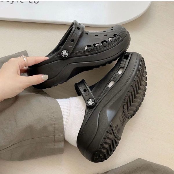 Black platform Crocs-style clogs with back strap, worn by a person, on a light wooden floor.