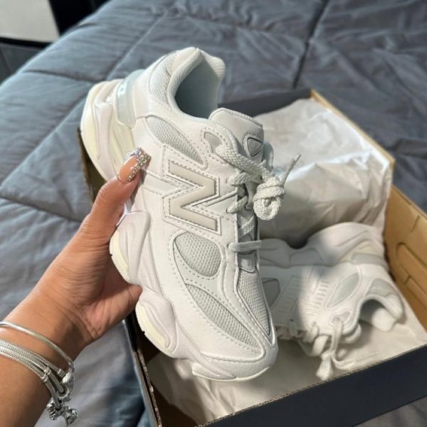 White chunky sneaker held in hand with another shoe and box visible in the background on a bed.