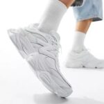 White athletic sneakers with chunky sole and mesh-panels, shown in a walking pose with white socks and light jeans.