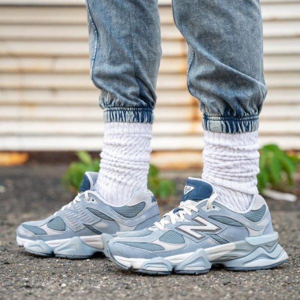 Close-up of legs wearing light-wash jeans, thick white scrunched socks, and gray/blue athletic sneakers standing on asphalt.