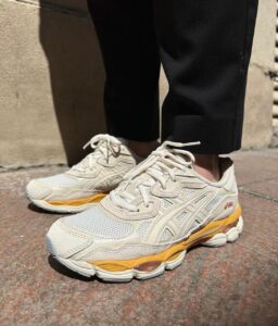 Close-up of white ASICS running sneakers with yellow midsole, worn by a person in black pants standing on stone pavement.