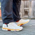 Close-up of white ASICS running shoes with orange and yellow sole details on a gray pavement, with dark pants visible and a wooden door in the background.