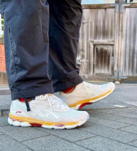 Close-up of white ASICS running shoes with orange and yellow sole details on a gray pavement, with dark pants visible and a wooden door in the background.