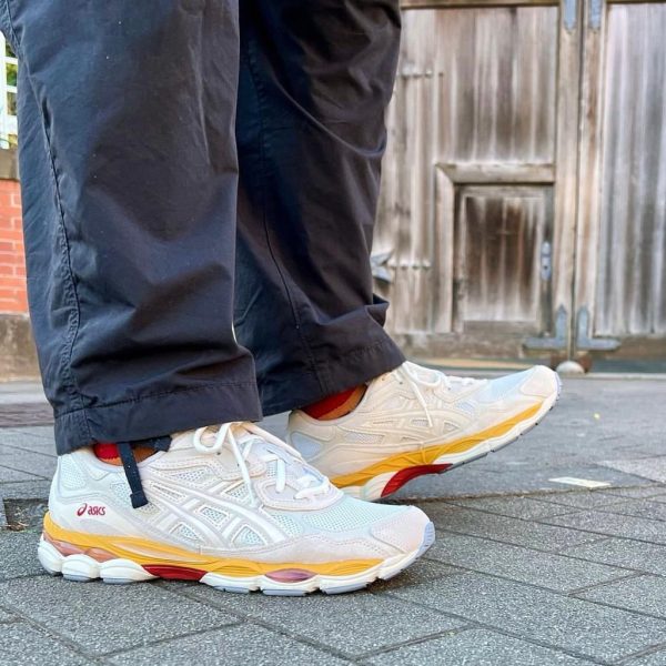 Close-up of white ASICS running shoes with orange and yellow sole details on a gray pavement, with dark pants visible and a wooden door in the background.