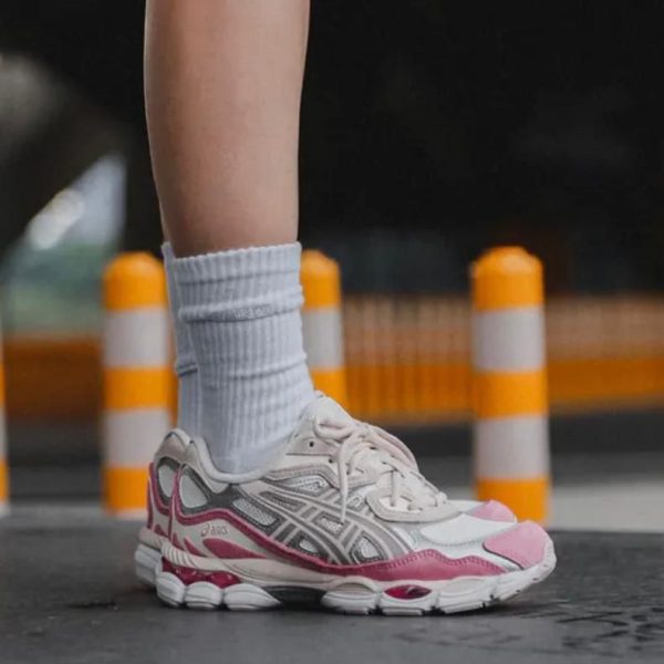 Close-up of a person wearing white and pink running shoes with white socks, standing on a street near orange-and-white traffic cones.
