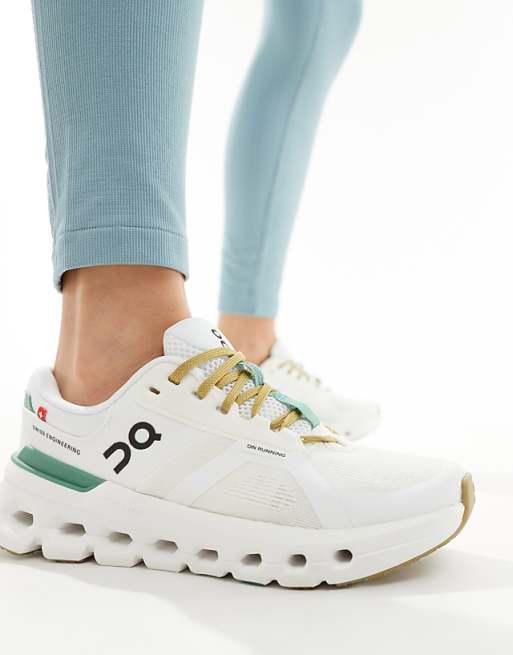 White running shoes with gold laces and a chunky sole on a person wearing light blue leggings; brand logo visible on the side.