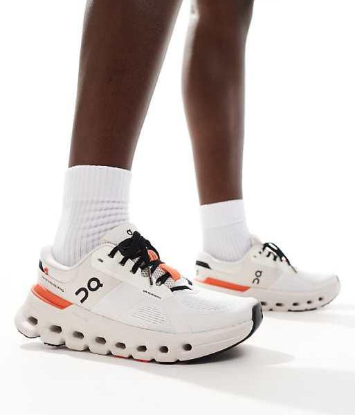 Person wearing white running shoes with a chunky sole and orange heel accents, standing with white socks.