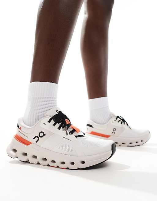 Person wearing white running shoes with a chunky sole and orange heel accents, standing with white socks.