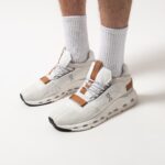 White athletic sneakers with tan-brown accents worn with light gray ribbed socks on a pale backdrop (showing a sporty lifestyle product).
