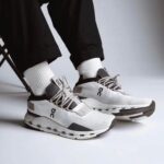 White and gray On-brand running shoes on a person wearing white socks and black pants, standing on a light surface.