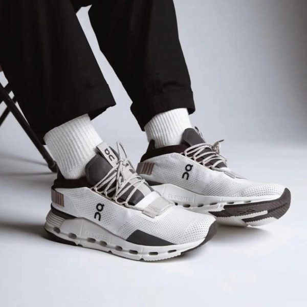 White and gray On-brand running shoes on a person wearing white socks and black pants, standing on a light surface.