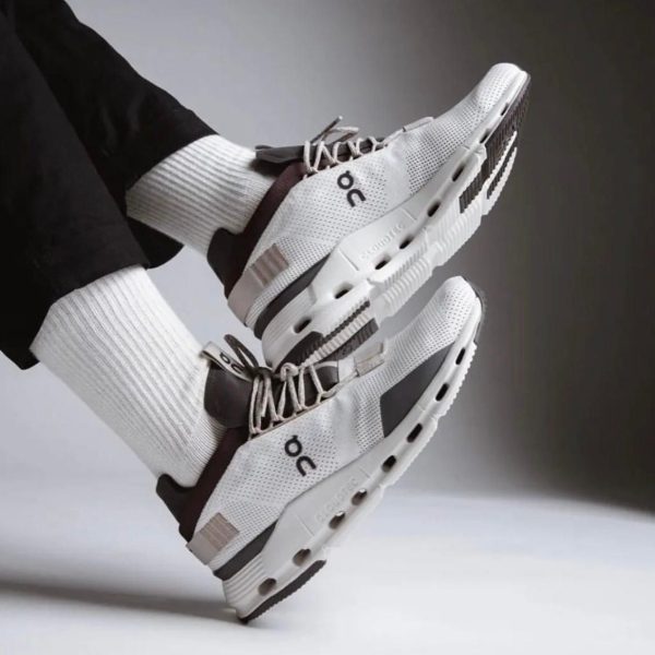White and gray running shoes with perforated knit upper, laces tied, and thick cushioned sole, worn with white socks by a person hovering their feet.