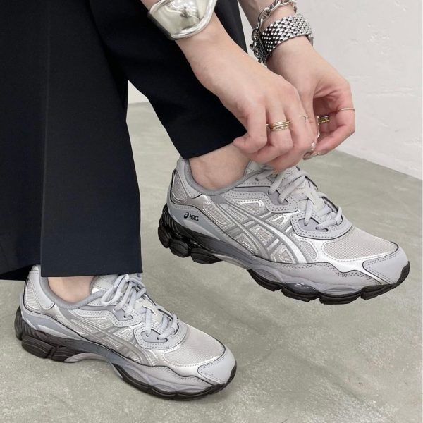 Person tying gray Asics running shoes, wearing multiple silver bracelets and rings, with black pants in a minimal setting.