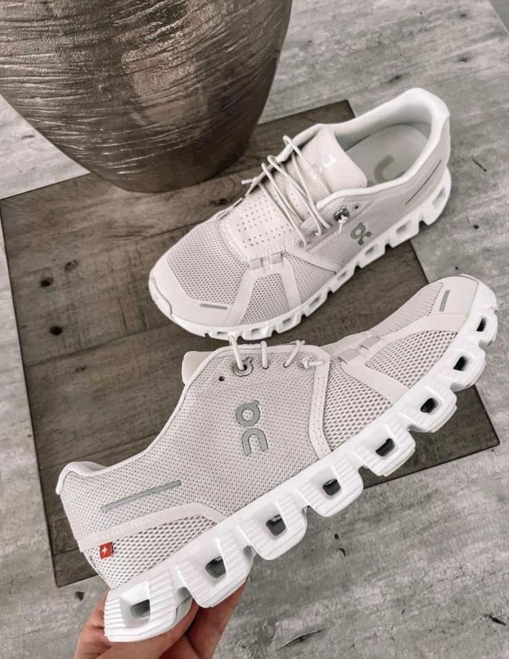 WhatsApp_Image_2024-11-10_at_13.42.15 Pair of light gray On running shoes with thick, segmented white soles on a wooden surface, showing the brand logo on the side.