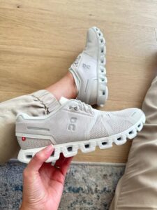 Gray mesh running shoe held in hand, showing rugged hollow sole and ON logo on the side, indoor wooden floor background.