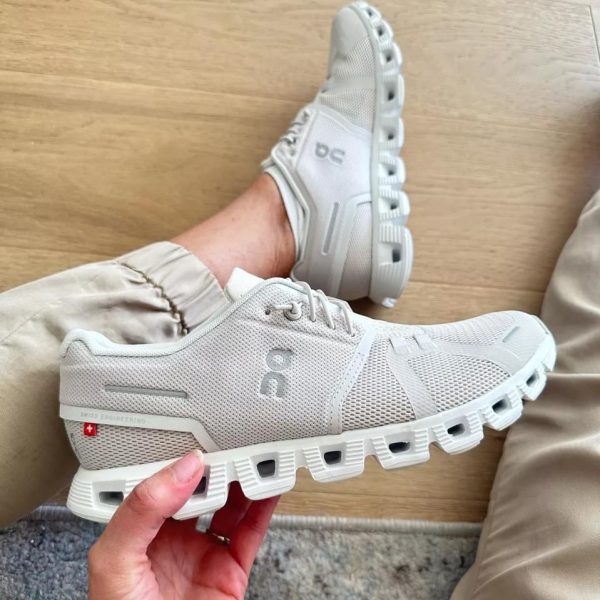 Gray mesh running shoe held in hand, showing rugged hollow sole and ON logo on the side, indoor wooden floor background.