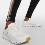 White athletic sneakers with perforated upper and chunky segmented sole, worn by someone in black leggings with a plaid side panel and an orange-tinted outsole at the toe area, shown in a side-view pose.