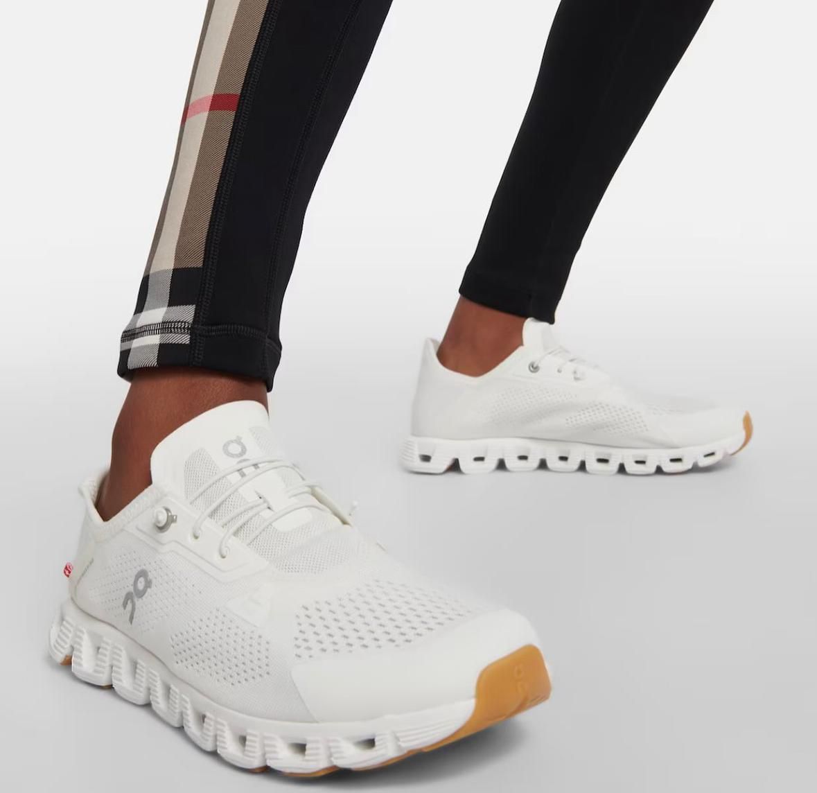 WhatsApp_Image_2024-11-11_at_13.13.42 White athletic sneakers with perforated upper and chunky segmented sole, worn by someone in black leggings with a plaid side panel and an orange-tinted outsole at the toe area, shown in a side-view pose.