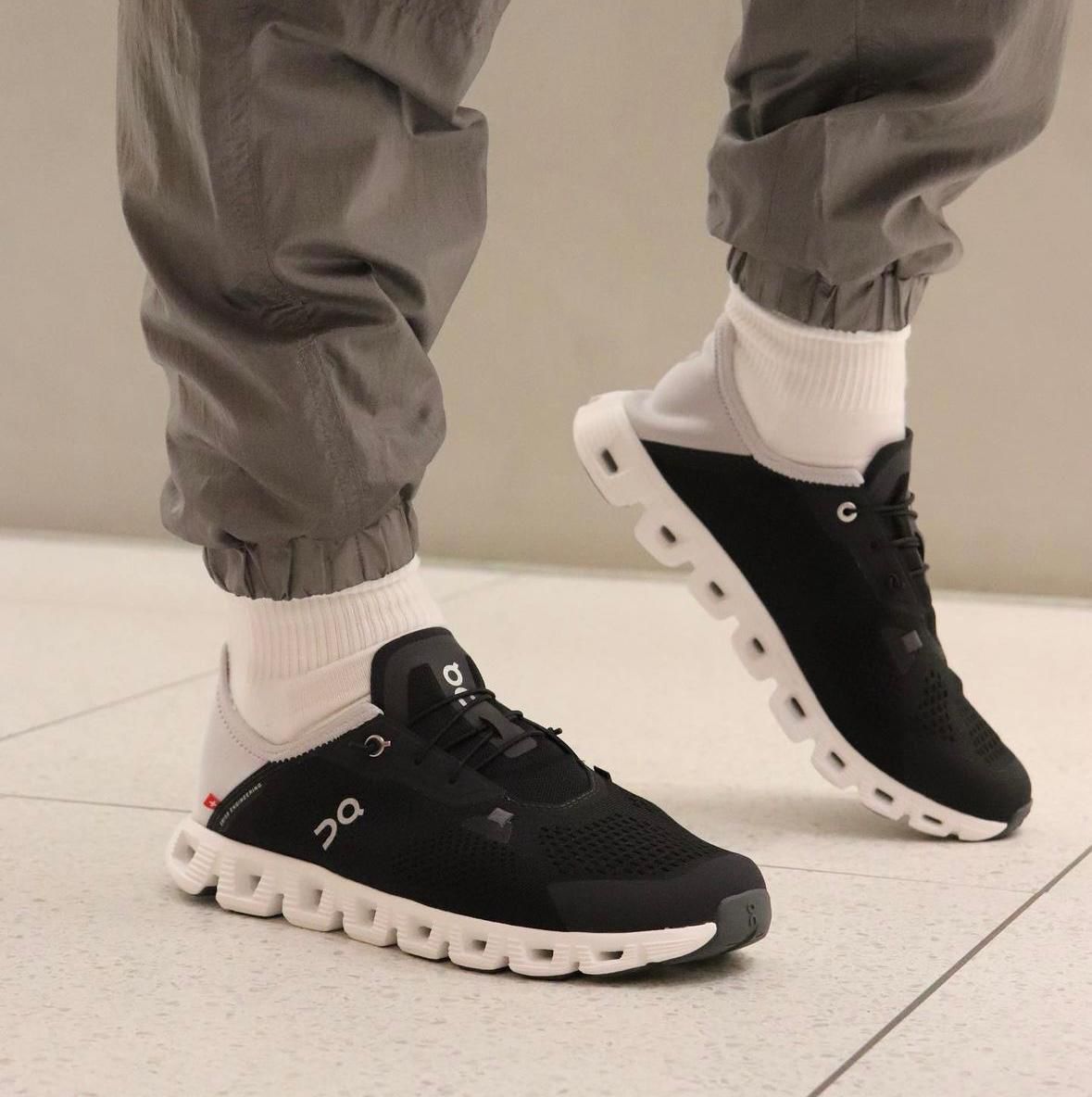 WhatsApp_Image_2024-11-11_at_14.04.25 Person wearing black and white athletic sneakers with chunky white sole, white socks, and gray joggers, mid-step on a tiled floor