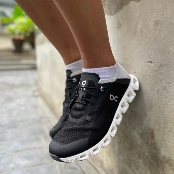 A person wearing black running shoes with a white chunky sole and white socks, leaning against a concrete wall.