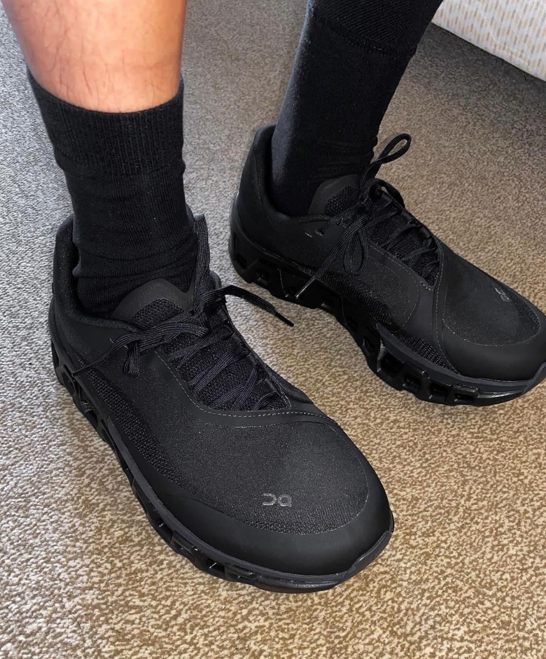 WhatsApp_Image_2024-11-12_at_14.02.52 | UAE SPORT STORE Person wearing black athletic sneakers and black socks standing on a carpeted floor, showcasing a chunky sole design.