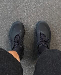 WhatsApp_Image_2024-11-12_at_14.03.02_1 | UAE SPORT STORE Top-down view of two black athletic sneakers on gray asphalt, seen from above. The laces are tied and the wearer’s legs are in dark pants.