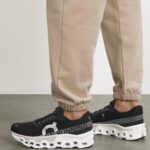 Beige joggers with black On running sneakers featuring a chunky white sole and visible cutouts at the bottom.