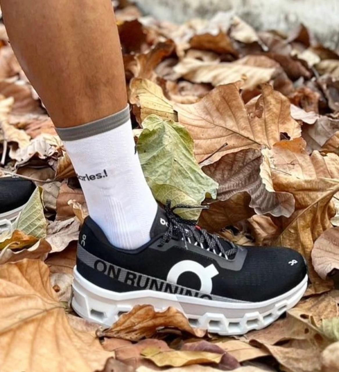 WhatsApp_Image_2024-11-13_at_13.44.21 | UAE SPORT STORE Close-up of a black On Running sneaker with a white sole and large logo, stepping on dry autumn leaves with a white sock nearby.