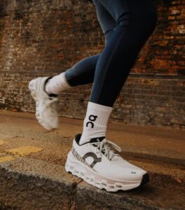 Close-up of a white athletic shoe stepping onto a curb, visible white sock with 'On' logo behind it, brick wall backdrop.