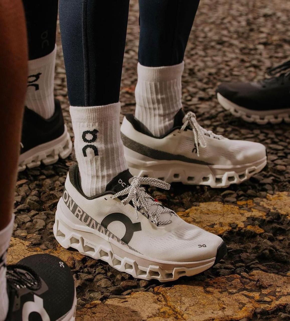 WhatsApp_Image_2024-11-13_at_15.46.57 Pair of white On running trail shoes with chunky built-up midsoles and white socks on a rocky gravel path.