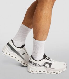 White running shoes with a perforated midsole and large hollowed-out sole, worn with light gray socks on a person’s feet, On logo visible on shoe side.