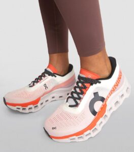 Pair of pink-white On running shoes with orange accents, worn with brown leggings, shown from the side/front profile.