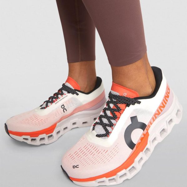 Pair of pink-white On running shoes with orange accents, worn with brown leggings, shown from the side/front profile.