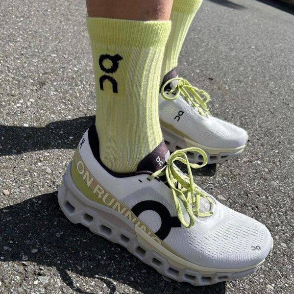 Person wearing white On running sneakers with lime green laces and bright yellow socks standing on asphalt road.