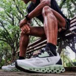 Close-up of a person tying the laces of white mint trail running shoes while sitting on a park bench outdoors.