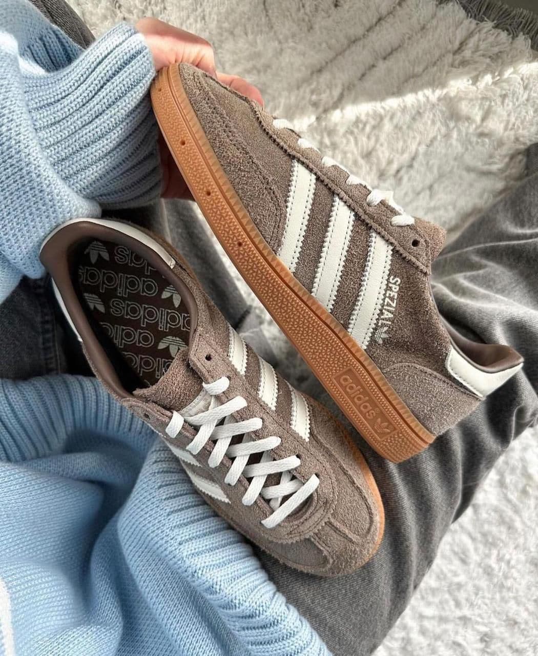 WhatsApp_Image_2024-11-22_at_11.10.56 | UAE SPORT STORE Brown suede Adidas sneakers with white stripes and gum sole held against a blue knit sweater.