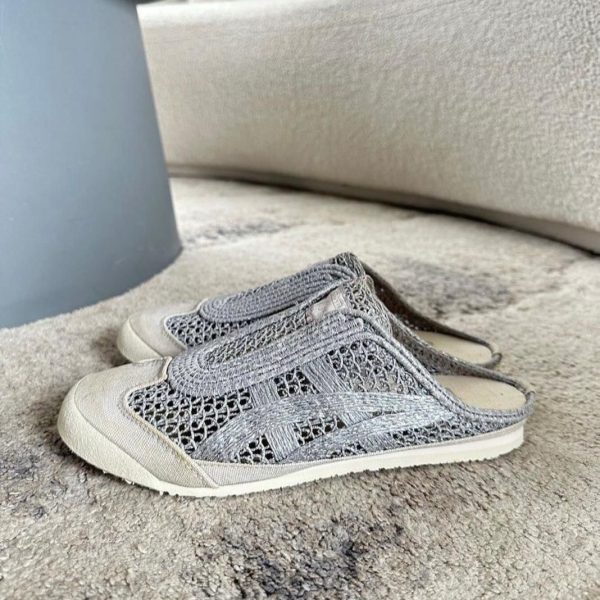 Pair of gray knitted slip-on sneakers with white soles resting on a light gray carpet near a gray furniture leg.