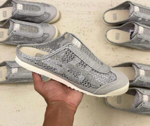 A hand holds up a silver woven sneaker with a light sole, surrounded by several matching shoes on a light floor.