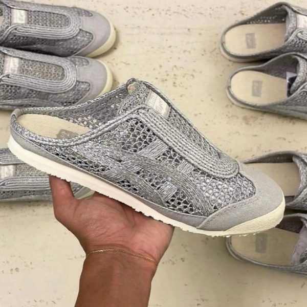 A hand holds up a silver woven sneaker with a light sole, surrounded by several matching shoes on a light floor.