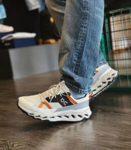 Close-up of a white and orange athletic sneaker on a person’s foot wearing blue jeans indoors on a wood floor, with a black side panel showing the logo.”] ,