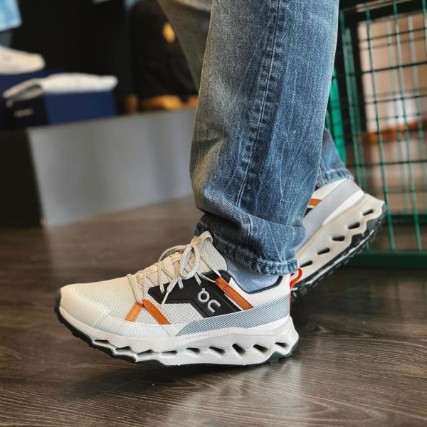 Close-up of a white and orange athletic sneaker on a person’s foot wearing blue jeans indoors on a wood floor, with a black side panel showing the logo.”] ,