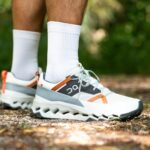 Pair of white and gray running shoes with orange accents on a trail, worn with white socks.