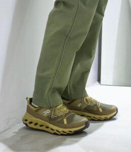 Legs in olive-green pants wearing tan trail-running shoes with a chunky, lattice-like sole (On brand).