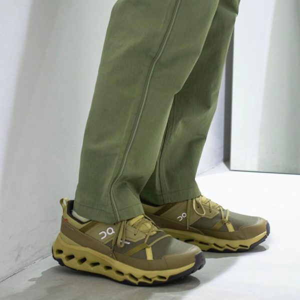 Legs in olive-green pants wearing tan trail-running shoes with a chunky, lattice-like sole (On brand).