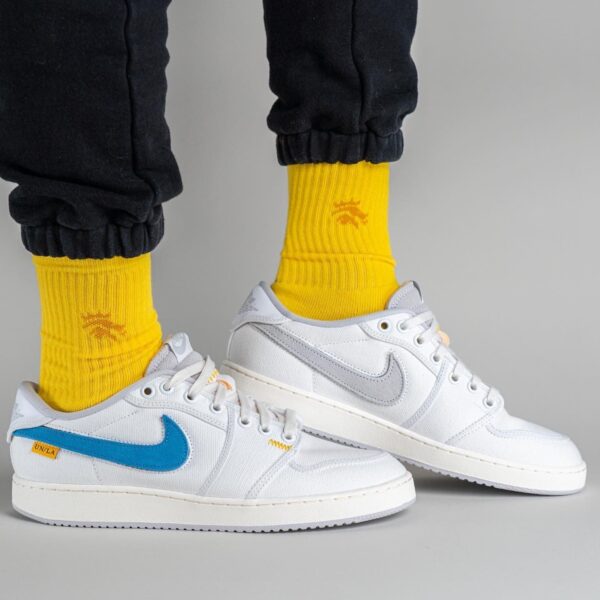 Close-up of feet in white Nike sneakers with a blue swoosh, paired with bright yellow socks and black joggers against a gray backdrop.