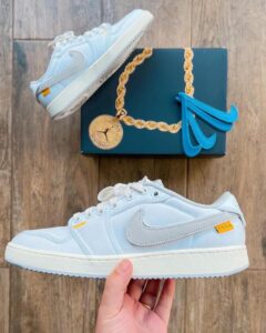 Light blue Nike sneakers held in a hand with a dark blue box and gold chain and blue swoosh accessories in the background.