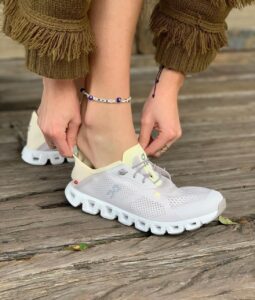 WhatsApp_Image_2024-12-14_at_14.17.03 | UAE SPORT STORE Person tying white athletic shoes on a wooden deck, wearing a fringed brown sweater and bracelets—casual outdoor scene.