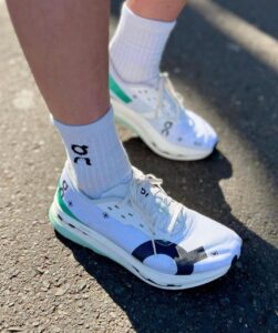 WhatsApp_Image_2024-12-15_at_10.40.47 | UAE SPORT STORE Person wearing white athletic sneakers and white socks standing on a paved surface, sunlight visible on the ground.